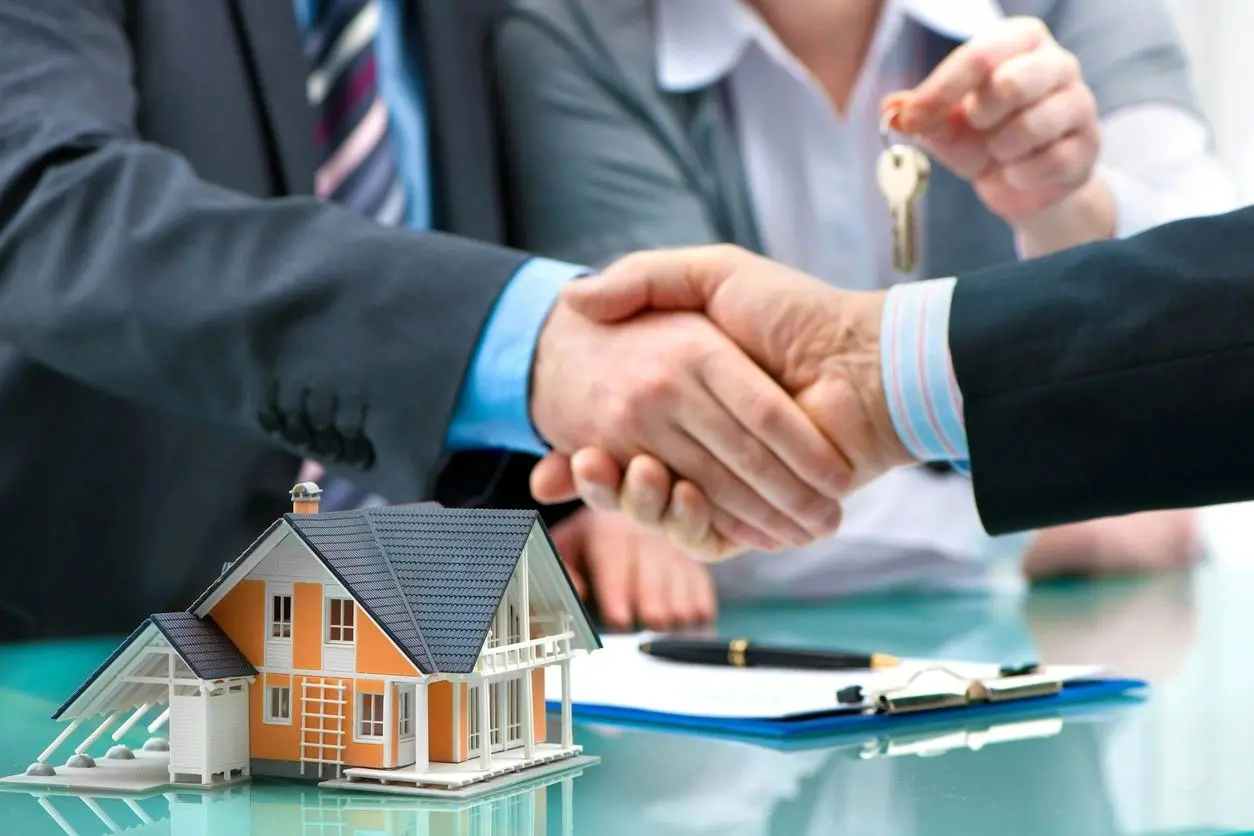 Two people shaking hands over a house model and some paperwork